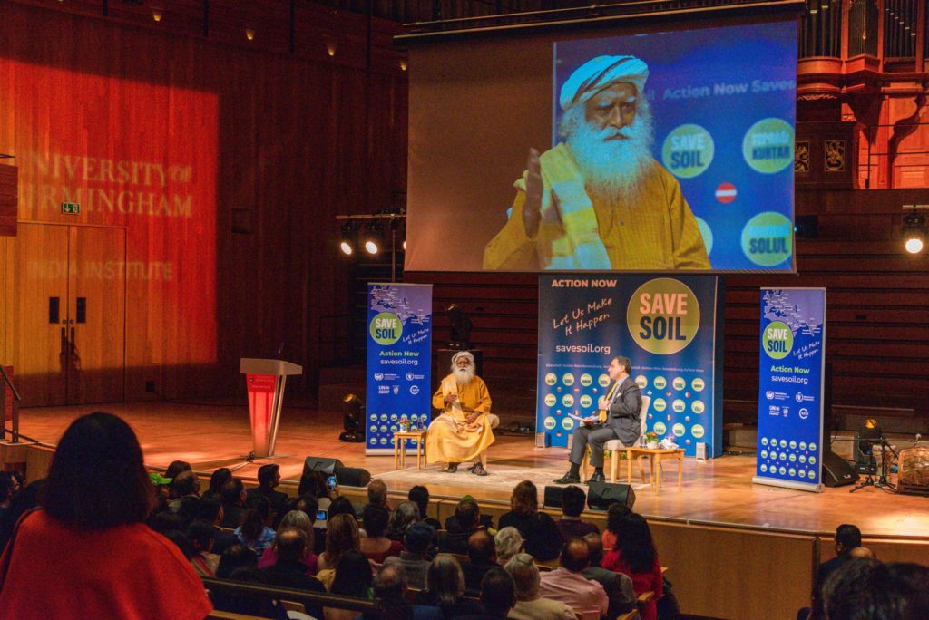 Sadhguru and Lord Karan Bilimoria in discussion on stage, with large projector screen overhead