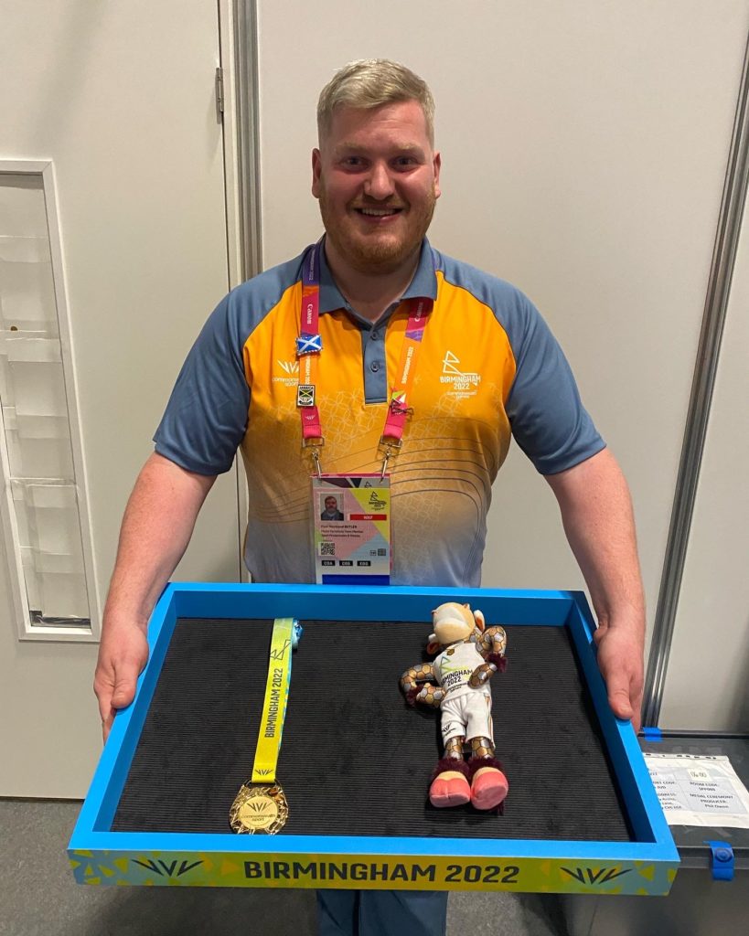 Smiling Paul in his grey and orange Birmingham 2022 volunteer uniform. Paul holds a tray with a gold medal and Perry on.