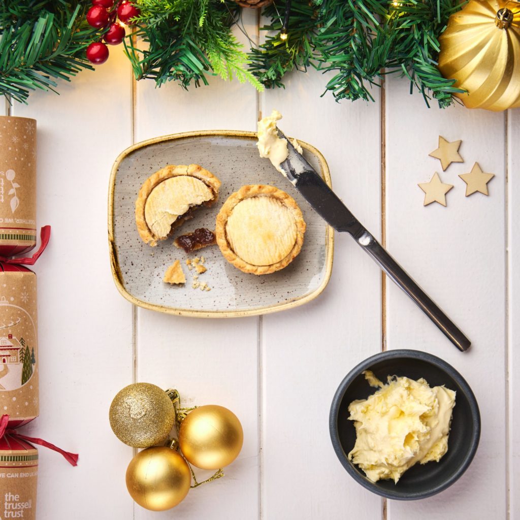 Mince pie on plate next to clotted cream. Christmas cracker and decorations frame the image.