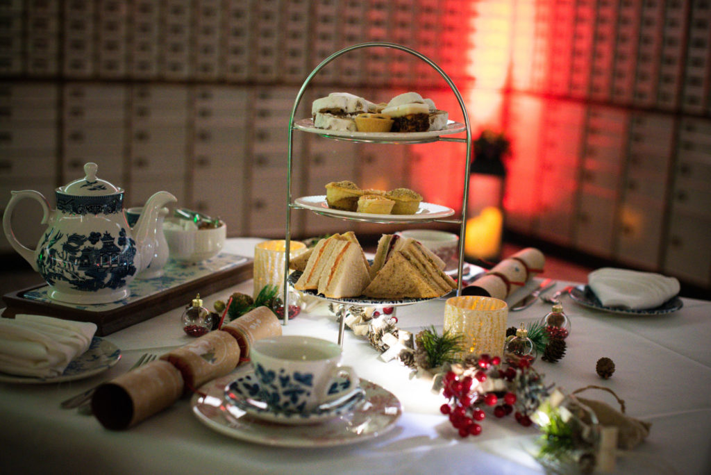 Table laid for a Christmas afternoon tea. Fine china is on the table, along with a stand with sandwiches and sweet treats,