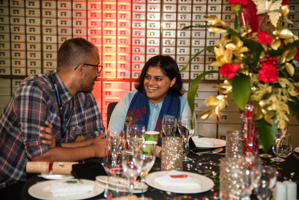 Two people sat at table laughing at Christmas party. Glitter and confetti are on the table and original bank vaults can be seen in the background.