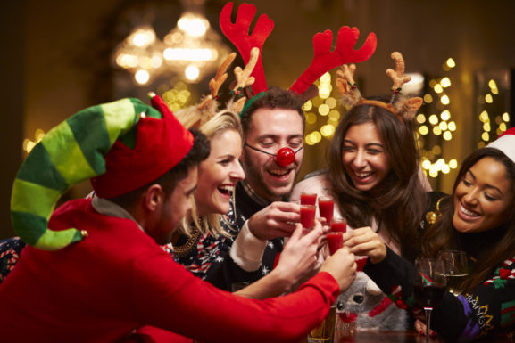 People at a Christmas party dressed in elf hats and reindeer antlers, toasting to the season with red shots.